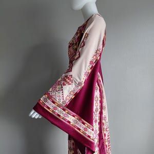 May include: A long, flowing dress with wide sleeves, displayed on a mannequin. The dress features a cream-colored bodice and sleeves, with a deep burgundy panel down the front. Intricate geometric patterns in red, yellow, and green adorn the sleeves and edges.