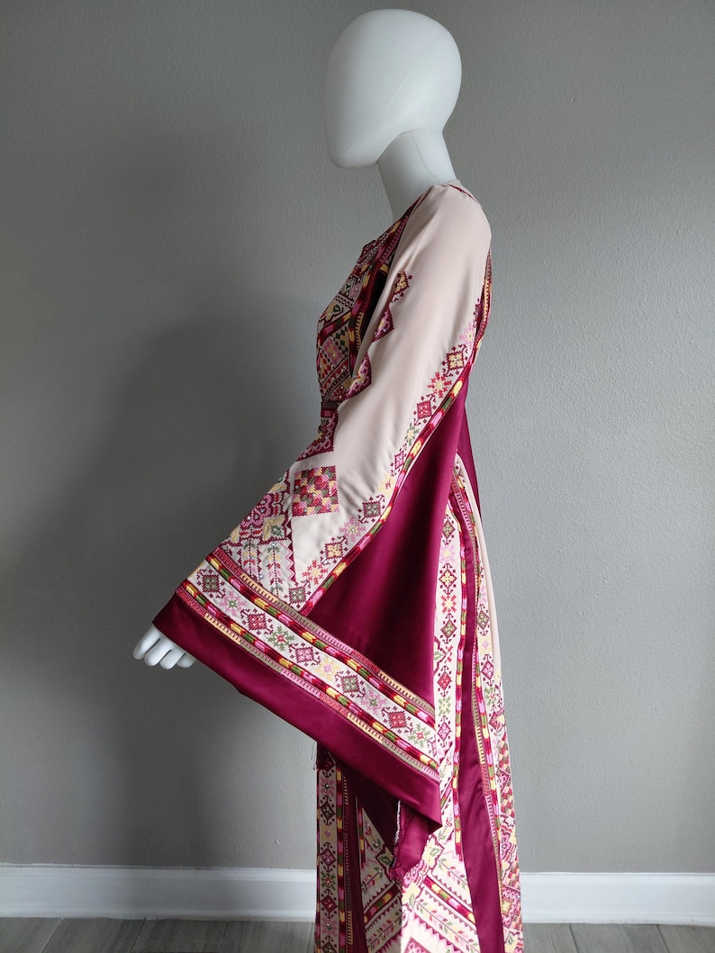 May include: A long, flowing dress with wide sleeves, displayed on a mannequin. The dress features a cream-colored bodice and sleeves, with a deep burgundy panel down the front. Intricate geometric patterns in red, yellow, and green adorn the sleeves and edges.