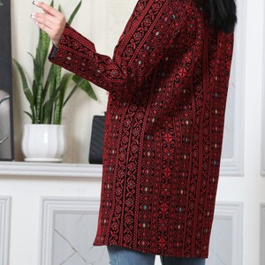 May include: A long, open-front jacket in a striking red and black pattern. The jacket features intricate embroidered details with a geometric design. The sleeves are long, and the jacket falls to mid-thigh. The jacket is worn over blue jeans.