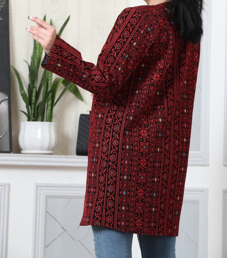 May include: A long, open-front jacket in a striking red and black pattern. The jacket features intricate embroidered details with a geometric design. The sleeves are long, and the jacket falls to mid-thigh. The jacket is worn over blue jeans.
