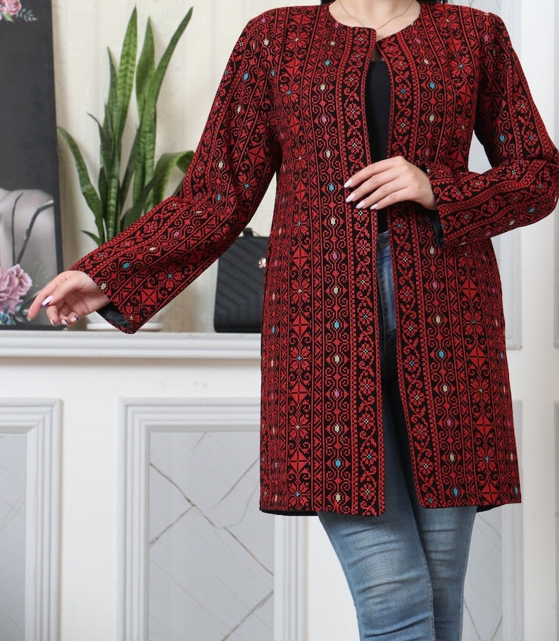 May include: A long, open-front red jacket with intricate black embroidery. The jacket features a detailed floral and geometric pattern, with accents of blue, green, and yellow. The sleeves are long, and the jacket falls to mid-thigh. The jacket is worn over a black top and blue jeans.