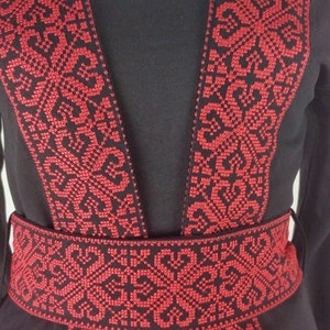 Puede incluir: Vestido negro con ribetes bordados en rojo alrededor del escote, las mangas y la cintura. El bordado presenta un patr&oacute;n geom&eacute;trico. El texto "Ramallah Embroidery" aparece dos veces en el vestido.