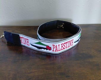 May include: White strap with the word "PALESTINE" in red, and a green, black, and red graphic. The strap has a black leather backing and gold-colored hardware. The strap is lying on a dark wooden surface.