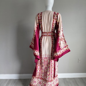 May include: A long, flowing dress with a cream-colored bodice and sleeves, accented with a deep burgundy trim and intricate geometric patterns in shades of red, yellow, and green. The dress features a matching belt and wide sleeves.