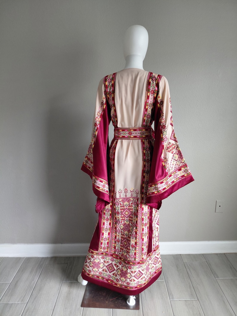 May include: A long, flowing dress with a cream-colored bodice and sleeves, accented with a deep burgundy trim and intricate geometric patterns in shades of red, yellow, and green. The dress features a matching belt and wide sleeves.