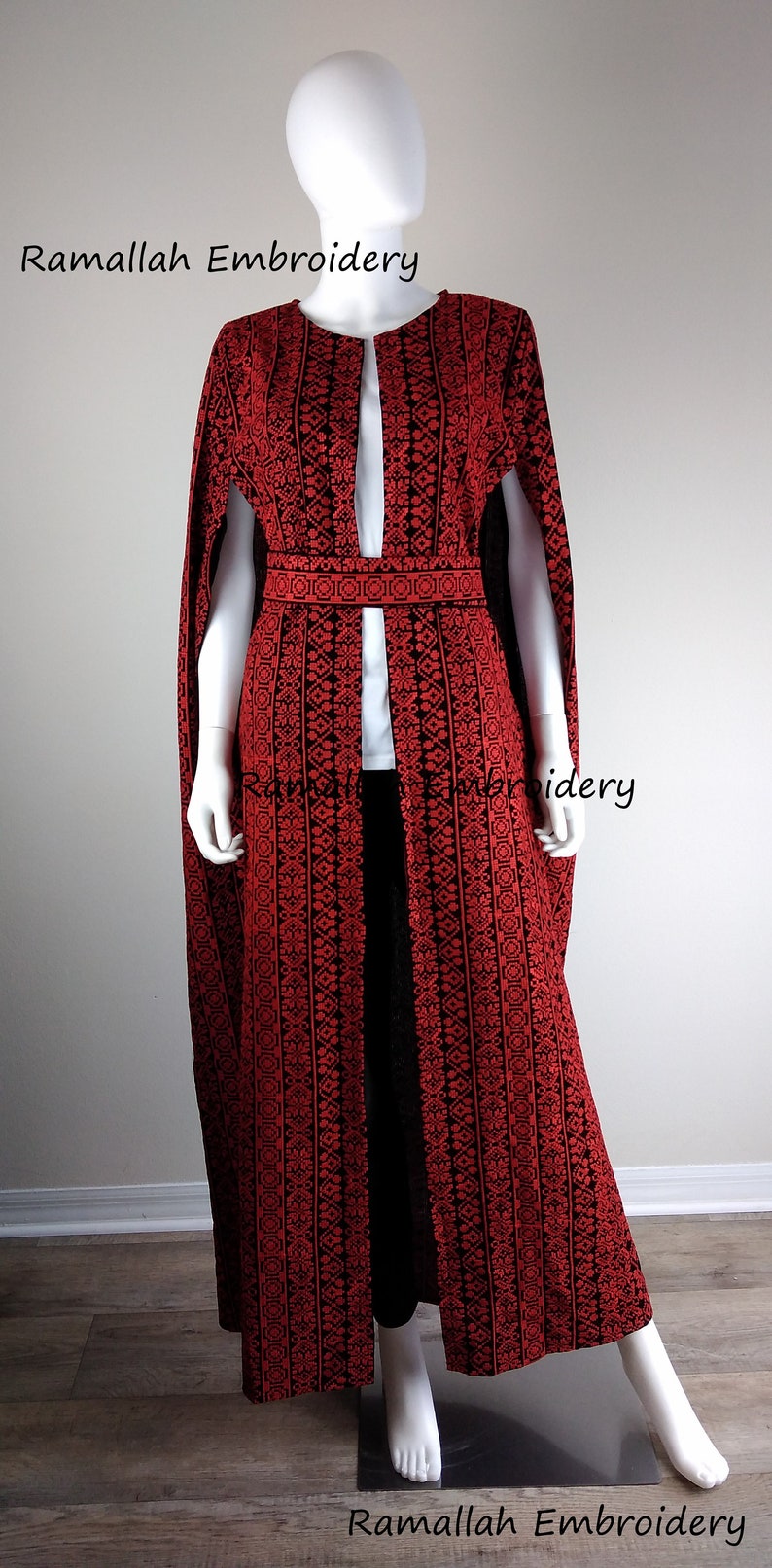 May include: A long, red embroidered cape with a black background and a black belt. The cape has a deep V-neck and is open in the front. The text "Ramallah Embroidery" is visible at the top and bottom of the image.