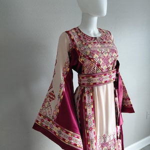 May include: A cream-colored dress with burgundy accents and intricate embroidery. The dress features long, flared sleeves and a matching belt. The bodice and sleeves are adorned with detailed patterns in shades of pink, yellow, and green.