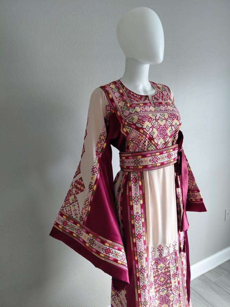May include: A cream-colored dress with burgundy accents and intricate embroidery. The dress features long, flared sleeves and a matching belt. The bodice and sleeves are adorned with detailed patterns in shades of pink, yellow, and green.