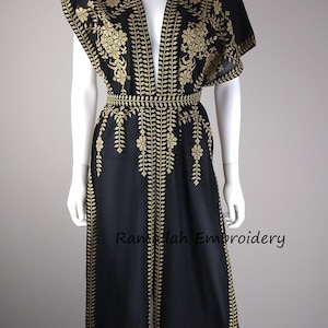 May include: Black embroidered jumpsuit with gold floral and leaf patterns. The open-front garment features a V-neck, short sleeves, and a belted waist. The text "Ramallah Embroidery" is visible.