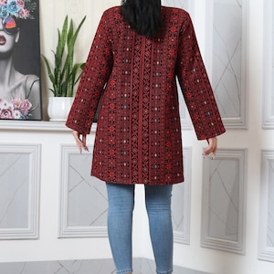 May include: A long-sleeved, red tunic with intricate black embroidery and colorful accents. The tunic is paired with light blue jeans. The overall style is a blend of traditional and modern fashion.