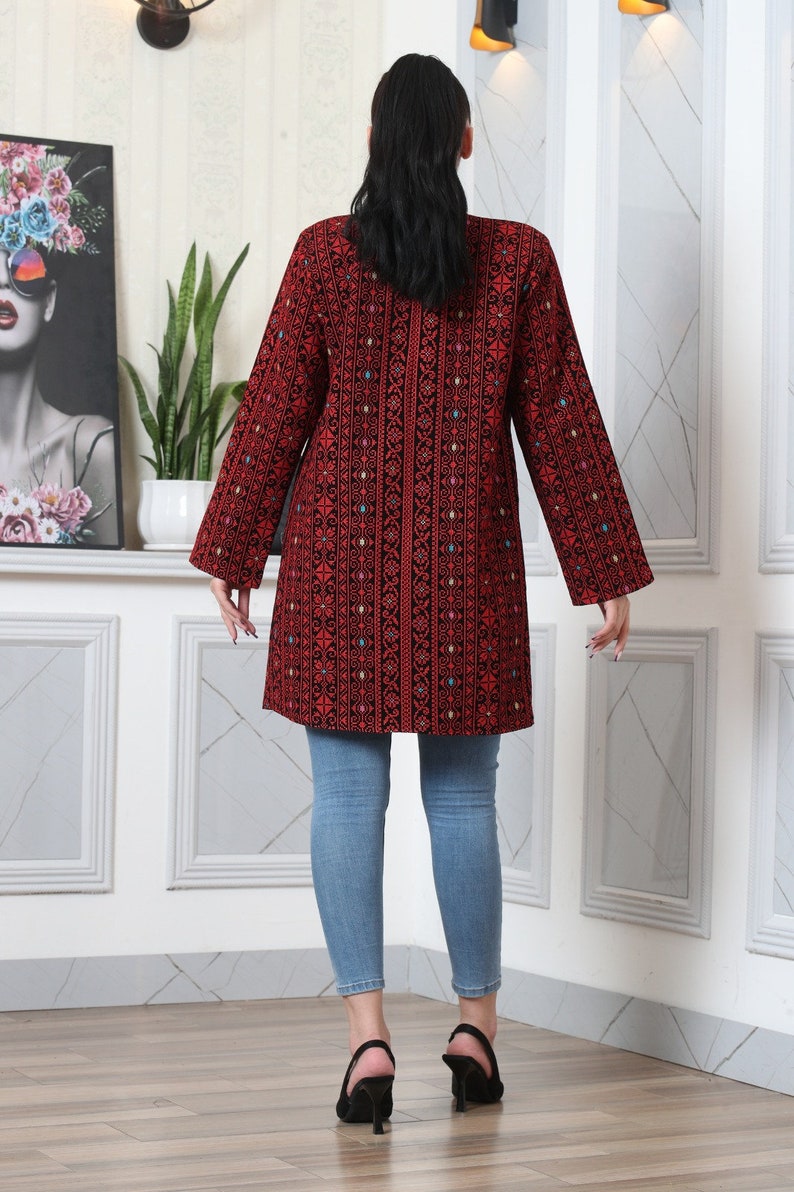 May include: A long-sleeved, red tunic with intricate black embroidery and colorful accents. The tunic is paired with light blue jeans. The overall style is a blend of traditional and modern fashion.
