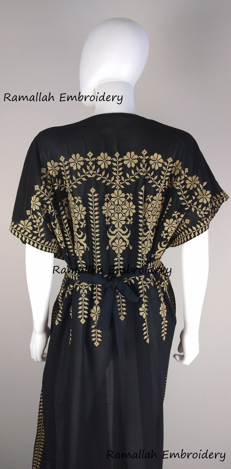 May include: Black kaftan dress with gold embroidery. The dress features a floral and leaf pattern across the shoulders and down the front. A matching tie belt cinches the waist. The text "Ramallah Embroidery" is visible.