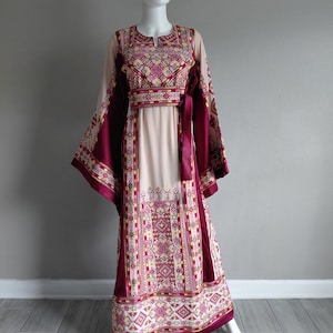 May include: A long, flowing dress on a mannequin. The dress features a cream-colored bodice and sleeves, with intricate embroidered patterns in burgundy, yellow, and pink. The dress has long, wide sleeves and a matching burgundy sash at the waist.