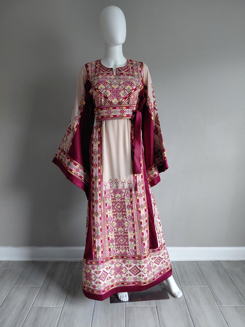 May include: A long, flowing dress on a mannequin. The dress features a cream-colored bodice and sleeves, with intricate embroidered patterns in burgundy, yellow, and pink. The dress has long, wide sleeves and a matching burgundy sash at the waist.