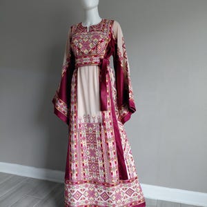 May include: A long, cream-colored dress with intricate embroidery in shades of burgundy, pink, and gold. The dress features a keyhole neckline, long sleeves, and a matching burgundy sash. The hemline and sleeves are adorned with detailed patterns.