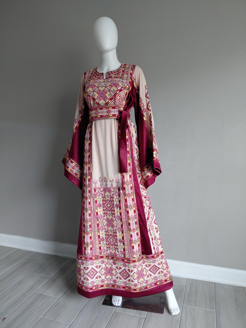 May include: A long, cream-colored dress with intricate embroidery in shades of burgundy, pink, and gold. The dress features a keyhole neckline, long sleeves, and a matching burgundy sash. The hemline and sleeves are adorned with detailed patterns.