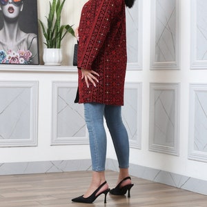 May include: A long, red coat with intricate black embroidery and a black fur collar. The coat is worn with light blue jeans and black heels. The coat's design features a detailed pattern, creating a visually rich garment.