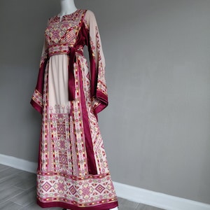 May include: A long, flowing dress with intricate embroidery. The dress features a cream-colored bodice and skirt, accented with burgundy panels and a matching belt. The sleeves are long and patterned, and the hemline is detailed with geometric designs.