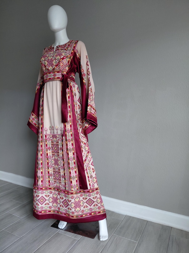 May include: A long, flowing dress with intricate embroidery. The dress features a cream-colored bodice and skirt, accented with burgundy panels and a matching belt. The sleeves are long and patterned, and the hemline is detailed with geometric designs.