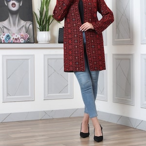 May include: A long, open-front red jacket with intricate black embroidery. The jacket features a detailed pattern and long sleeves. The person is wearing blue jeans and black heels. The background includes a framed artwork and a white wall.