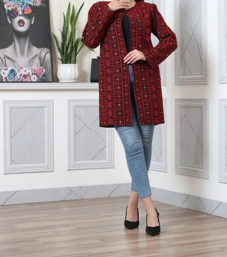 May include: A long, open-front red jacket with intricate black embroidery. The jacket features a detailed pattern and long sleeves. The person is wearing blue jeans and black heels. The background includes a framed artwork and a white wall.