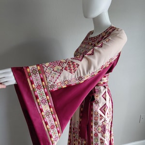May include: A long, flowing dress with wide sleeves. The dress features a cream-colored bodice and sleeves with intricate embroidered patterns in shades of red, pink, and green. The sleeves and skirt are a deep burgundy color, and a matching belt cinches the waist.