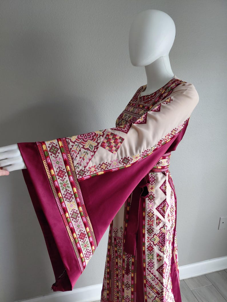 May include: A long, flowing dress with wide sleeves. The dress features a cream-colored bodice and sleeves with intricate embroidered patterns in shades of red, pink, and green. The sleeves and skirt are a deep burgundy color, and a matching belt cinches the waist.