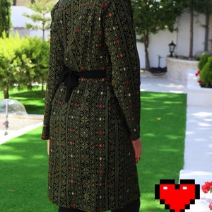 May include: A long, green and black patterned dress with a black belt. The dress has a traditional embroidered design with red and white accents.