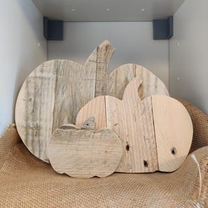Puede incluir: Tres decoraciones de calabazas de madera de diferentes tama&ntilde;os. Las calabazas est&aacute;n hechas de madera clara y tienen un aspecto r&uacute;stico y envejecido. Est&aacute;n dispuestas sobre una superficie de arpillera, creando una exhibici&oacute;n con tem&aacute;tica oto&ntilde;al.