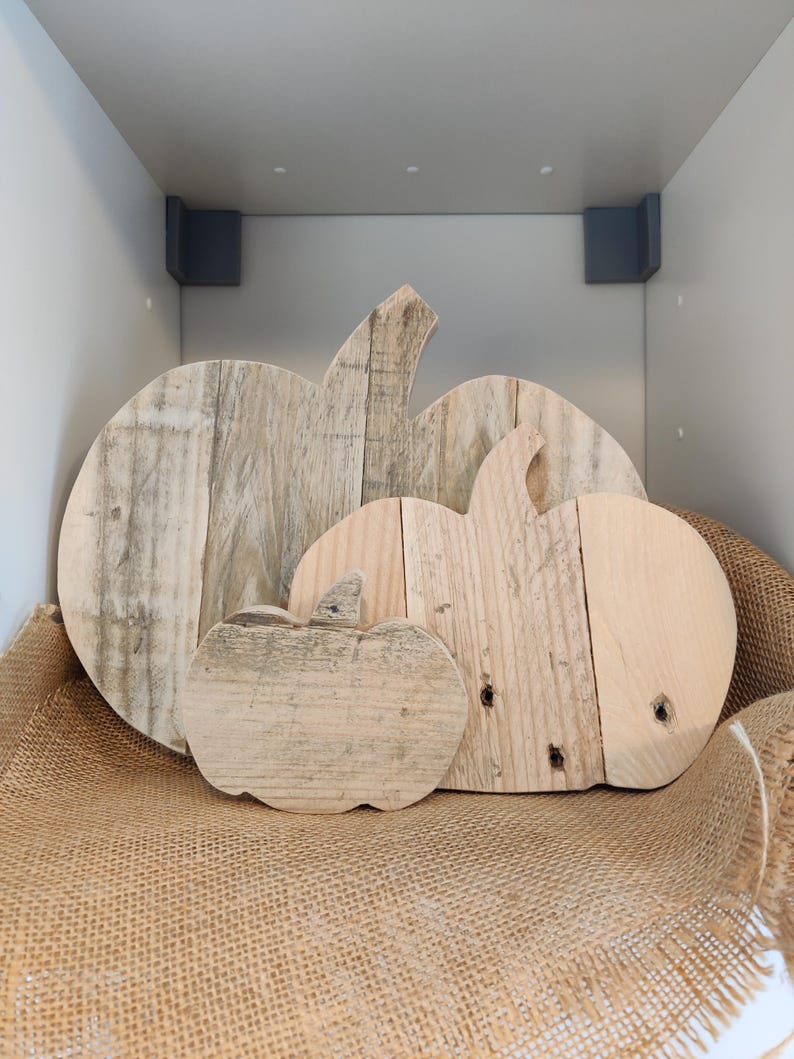 Puede incluir: Tres decoraciones de calabazas de madera de diferentes tama&ntilde;os. Las calabazas est&aacute;n hechas de madera clara y tienen un aspecto r&uacute;stico y envejecido. Est&aacute;n dispuestas sobre una superficie de arpillera, creando una exhibici&oacute;n con tem&aacute;tica oto&ntilde;al.