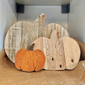 Puede incluir: Tres decoraciones de calabaza de madera de diferentes tama&ntilde;os. La calabaza m&aacute;s grande y la del medio son de madera clara, mientras que la m&aacute;s peque&ntilde;a est&aacute; pintada de naranja con un tallo marr&oacute;n. Las calabazas se exhiben sobre una superficie de arpillera.