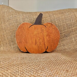 Puede incluir: Decoraci&oacute;n de calabaza de madera con cuerpo naranja y tallo marr&oacute;n oscuro. La calabaza est&aacute; colocada sobre un fondo de arpillera, creando un tema oto&ntilde;al r&uacute;stico. La superficie de la calabaza tiene vetas de madera visibles.