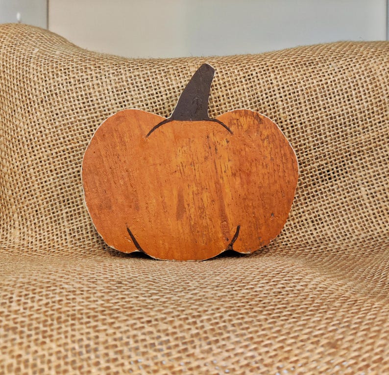 Puede incluir: Decoraci&oacute;n de calabaza de madera con cuerpo naranja y tallo marr&oacute;n oscuro. La calabaza est&aacute; colocada sobre un fondo de arpillera, creando un tema oto&ntilde;al r&uacute;stico. La superficie de la calabaza tiene vetas de madera visibles.