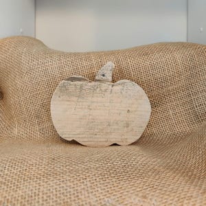 Puede incluir: Una decoraci&oacute;n r&uacute;stica de calabaza de madera con un acabado claro y desgastado. La calabaza tiene un tallo y est&aacute; colocada sobre un fondo de arpillera, evocando un tema de oto&ntilde;o o cosecha. La veta de la madera es visible.
