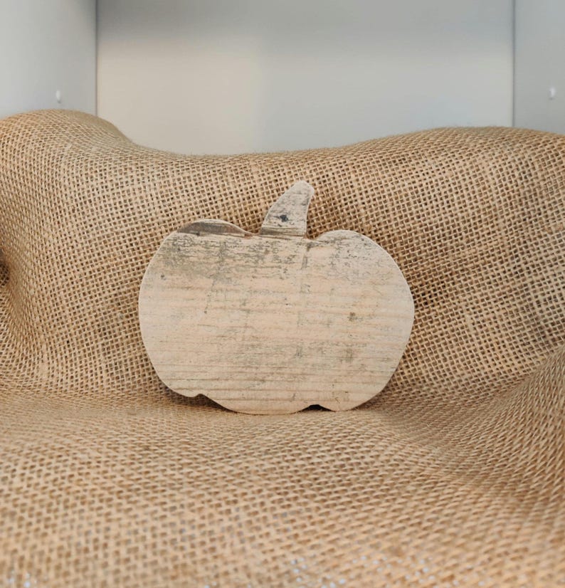 Puede incluir: Una decoraci&oacute;n r&uacute;stica de calabaza de madera con un acabado claro y desgastado. La calabaza tiene un tallo y est&aacute; colocada sobre un fondo de arpillera, evocando un tema de oto&ntilde;o o cosecha. La veta de la madera es visible.
