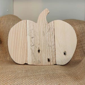 Puede incluir: Una decoraci&oacute;n de calabaza de madera hecha de tablas de madera de color claro. La calabaza tiene un tallo y se muestra sobre una superficie de arpillera. La veta de la madera es visible y la forma de la calabaza est&aacute; claramente definida.