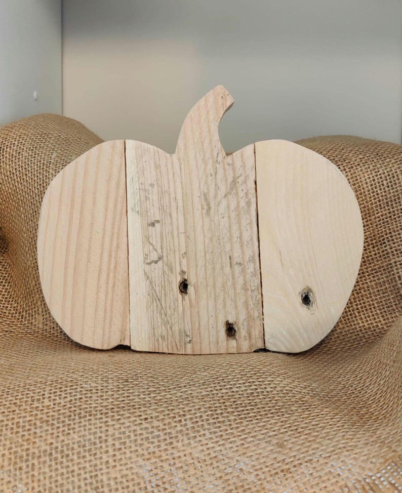Puede incluir: Una decoraci&oacute;n de calabaza de madera hecha de tablas de madera de color claro. La calabaza tiene un tallo y se muestra sobre una superficie de arpillera. La veta de la madera es visible y la forma de la calabaza est&aacute; claramente definida.