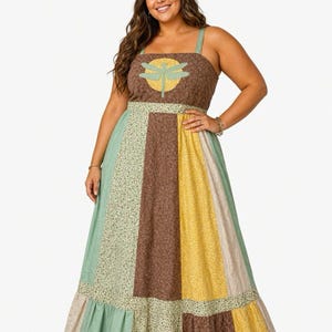 May include: A sleeveless maxi dress featuring a patchwork design with panels of brown, yellow, and sage green floral fabric. The bodice displays a yellow circular sun motif with a light green dragonfly graphic centered on the chest area.