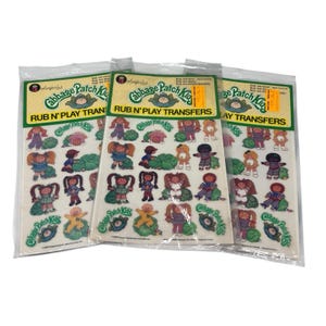 May include: Three sealed packages of Cabbage Patch Kids Rub N' Play Transfers. Each package features a yellow and green header with the Cabbage Patch Kids logo and various transfer designs of the dolls. The transfers are in a clear plastic packaging.