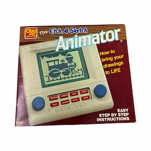 May include: The Ohio Art Etch A Sketch Animator features a beige drawing device with two blue knobs and red buttons labeled ON/MOVE, ANIMATE, SAVE, DRAW, NEXT, REVERSE, RECALL, and ERASE. The screen displays a pixelated train graphic on the box.