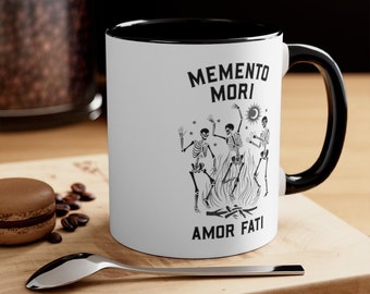 May include: White ceramic mug with a black rim and handle. The mug features a black and white illustration of three skeletons dancing around a fire with the text "Memento Mori Amor Fati".