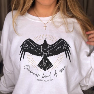 K&ouml;nnte beinhalten: Wei&szlig;er Pullover mit einer schwarzen Silhouette eines Vogels mit dem Text "Ominous bird of yore" und "Edgar Allan Poe" auf der Vorderseite.