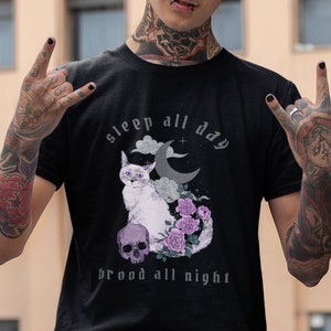K&ouml;nnte beinhalten: Ein schwarzes T-Shirt mit einem Motiv einer Katze, Mond und Blumen. Der Text auf dem Shirt lautet "sleep all day brood all night".