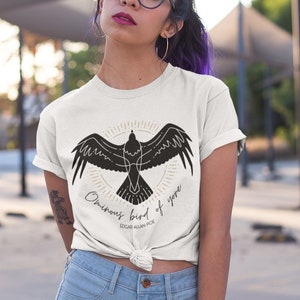 Puede incluir: Una camiseta blanca con una silueta negra de un p&aacute;jaro con el texto "Omniscient bird of yore" y "Edgar Allan Poe" debajo.