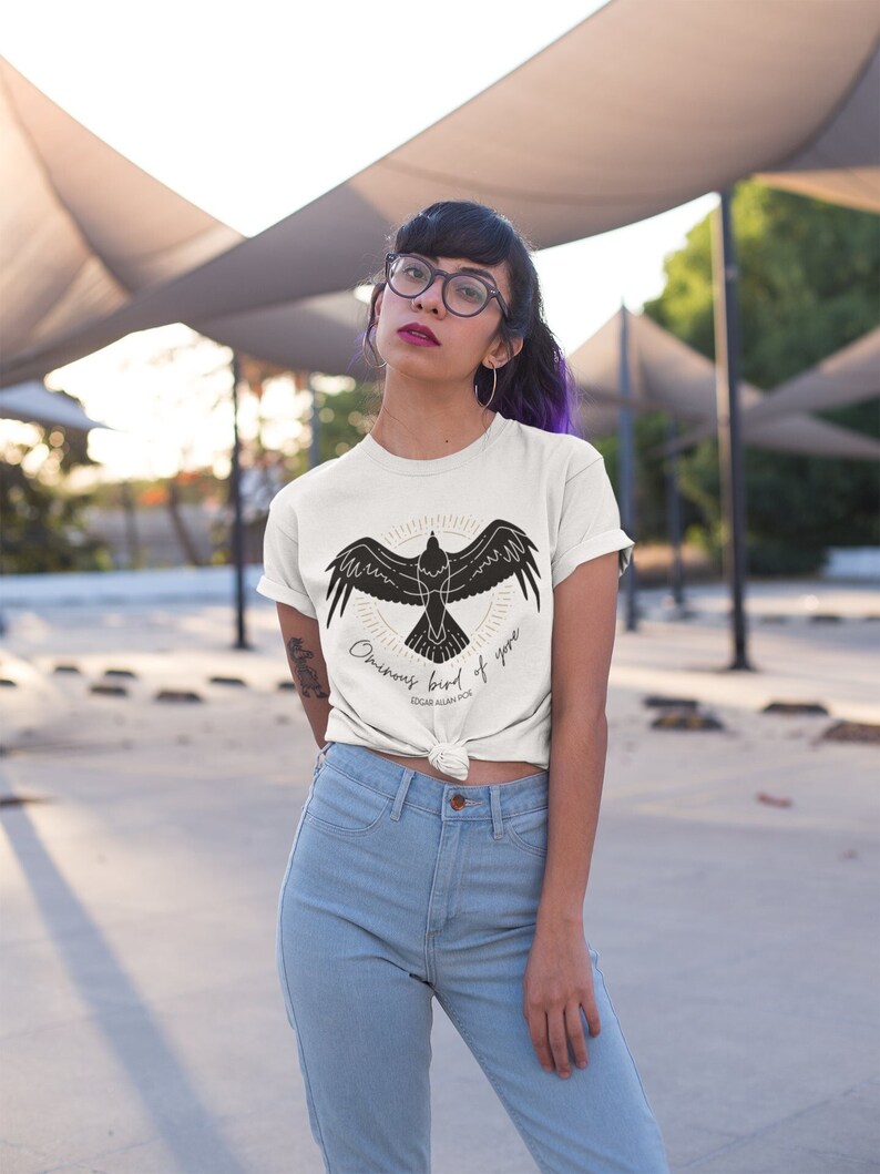 Puede incluir: Una camiseta blanca con una silueta negra de un p&aacute;jaro con el texto "Omniscient bird of yore" y "Edgar Allan Poe" debajo.