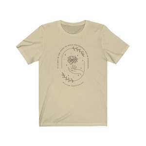 May include: A beige t-shirt with a quote from William Shakespeare: "It is not in the stars to hold our destiny but in ourselves." The quote is surrounded by a floral design with a hand holding a flower.