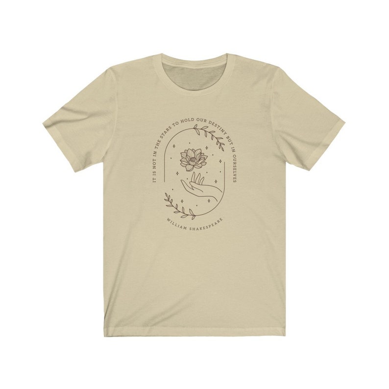 May include: A beige t-shirt with a quote from William Shakespeare: "It is not in the stars to hold our destiny but in ourselves." The quote is surrounded by a floral design with a hand holding a flower.
