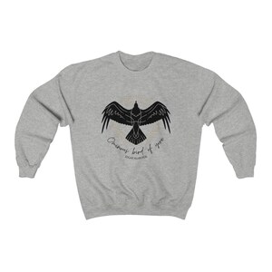 K&ouml;nnte beinhalten: Ein graues Sweatshirt mit einer schwarzen Silhouette eines Rabens im Flug und dem Text "Ominous bird of yore" und "Edgar Allan Poe" darunter.