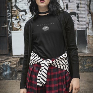 Puede incluir: Una mujer lleva un c&aacute;rdigan negro de manga larga sobre una camiseta negra con el texto "eat the rich" y un gr&aacute;fico de una boca con colmillos. Lleva una falda de cuadros rojos y negros con un cintur&oacute;n a rayas blancas y negras atado a la cintura.