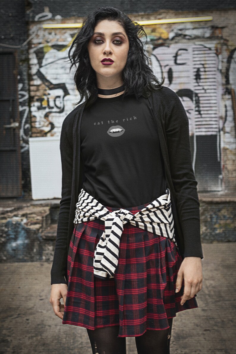 Puede incluir: Una mujer lleva un c&aacute;rdigan negro de manga larga sobre una camiseta negra con el texto "eat the rich" y un gr&aacute;fico de una boca con colmillos. Lleva una falda de cuadros rojos y negros con un cintur&oacute;n a rayas blancas y negras atado a la cintura.